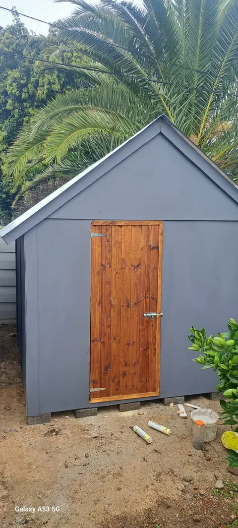 A grey timber toolshed with a stained timber door, on a bare site with a palm tree behind